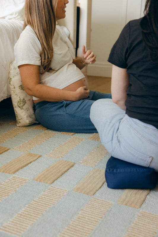Two pregnant women sitting together and talking; a back-view showing The Tush Present used as a seat for pelvic support and another as lumbar support for neutral spine alignment.