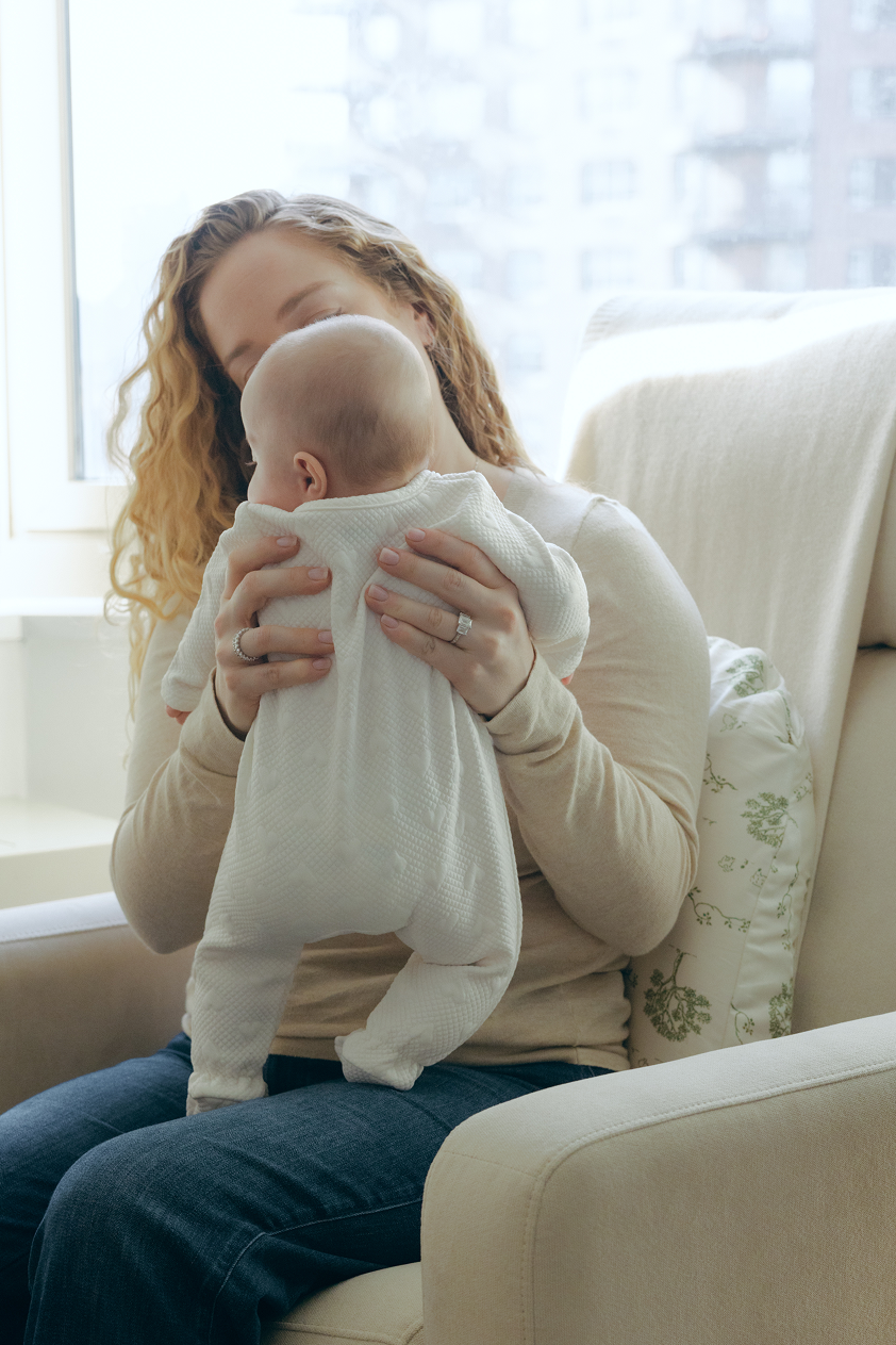 Woman holding a baby in a living room with a large window sitting against a tush pillow in a chair